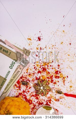 Top View Of Graceful Rakhi, Sweet And Indian Currency With Spread Rice Grains, Kumkum And Haldi On W