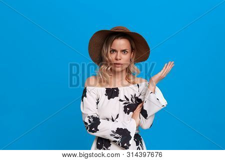Grumpy Young Model With Curly Blond Hair In A Sundown Hat And White Dress Looks At The Camera Standi