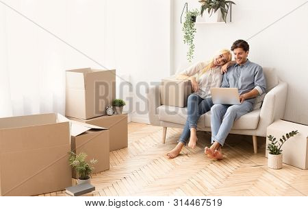 Designing New House. Cheerful Couple Relaxing On Couch With Laptop Among Moving Boxes. Panorama With