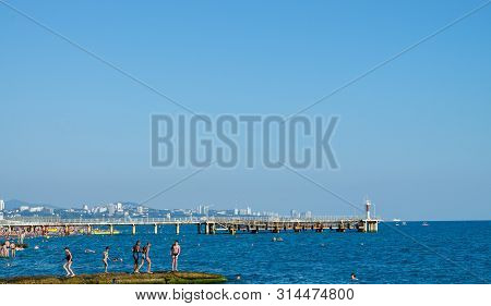 Sochi, Russia - July 7, 2019: People On The Breakwater Of Dagomys Beach Overlooking The Pier And Cit