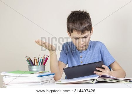 Confused, Surprised Child With Tablet Computer Sitting At Table With Books Notebooks