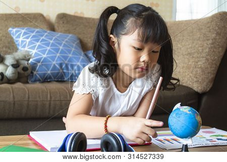 Asian Little Girl Playing With Globe On Table, Holding A Pencil In Her Hand Before Write In Notebook