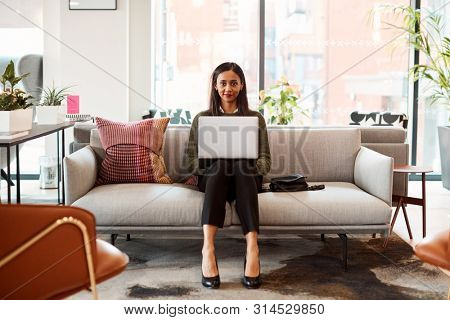 Portrait Of Businesswoman Sitting On Sofa Working On Laptop At Desk In Shared Workspace Office