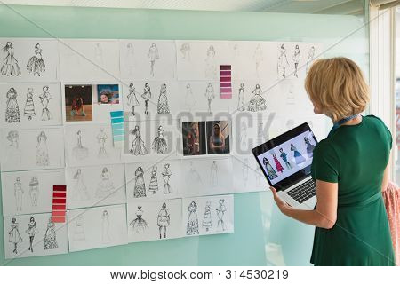 Rear view of beautiful mature Caucasian female fashion designer looking at sketch while holding laptop in design studio. Modern casual creative business concept
