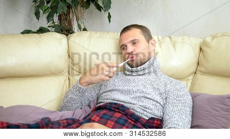 Young Sick Man In A Sweater Under A Plaid With A Fever Checks His Temperature With A Thermometer At 