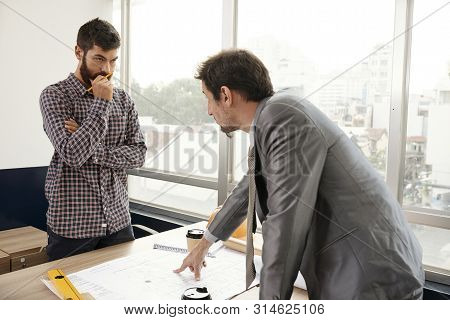 Businessman In Suit Standing Near The Table Pointing At Blueprint And Discussing Some Working Moment
