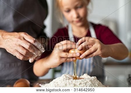 Closeup hands of child and senior woman breaking egg into flour for dough. Little girl wearing apron preparing pie with egg. Grandmother and granddaughter preparing dough for biscuits in kitchen.