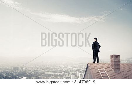 Young Determined Businessman Standing On House Roof And Looking Away. Mixed Media
