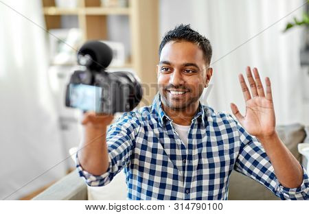 blogging, videoblog and people concept - smiling indian male video blogger with camera videoblogging and waving hand at home