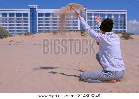 Woman Traveller On The Beach Throws Sand That Flies In The Wind. She Rests Sitting At Sea On Sand Du