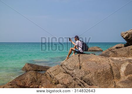 Man Talks Video Chat At Sea On Smartphone With Small Tripod Sits On Stone. Young Guy Traveller Share