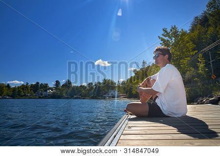 Young Guy Awaits Sitting On The Pier Ferry To Lake Massawippi, The Eastern City Of Magog, Quebec, La