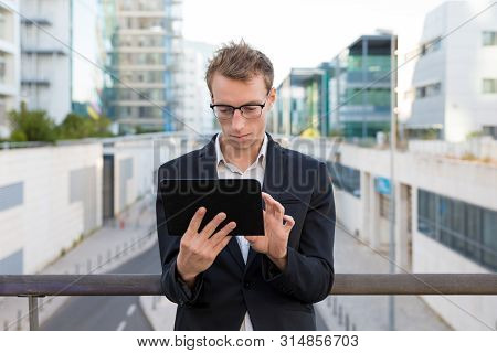 Focused Serious Manager Checking Messages On Tablet. Young Business Man In Eyeglasses Standing On Ov