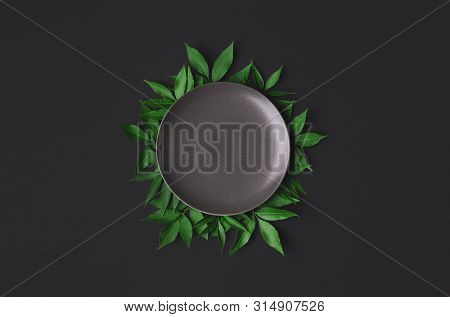 Empty Grey Plate On Black Background, Surrounded By Green Leaves. Flat Lay Of Empty Dish. Food Mocku