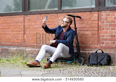 Smiling Businessman Sitting On Electric Scooter Near Brick Wall Taking Selfie With Mobile Phone