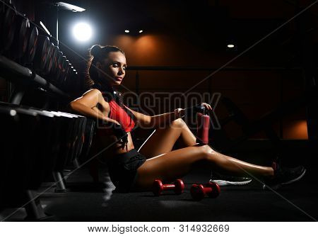 Pleased Satisfied With Herself Sporty Girl Is Sitting On The Floor At Stand With Dumbbells With Her 