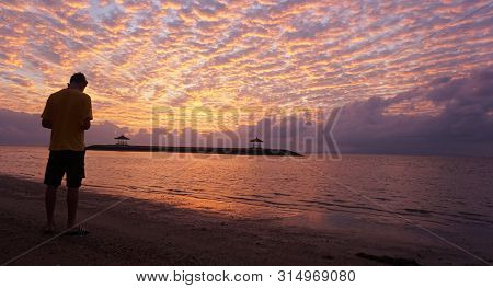 Young Man Standing With Head Down Against Morning Glow Sunrise Sky. Dramatic Colorful Sky And Beach 