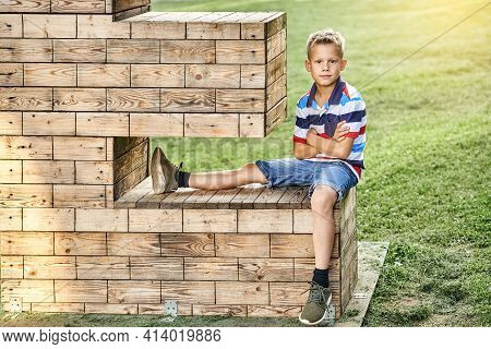 Little Boy In Polo T-shirt And Shorts Sits On Wooden Art Installation