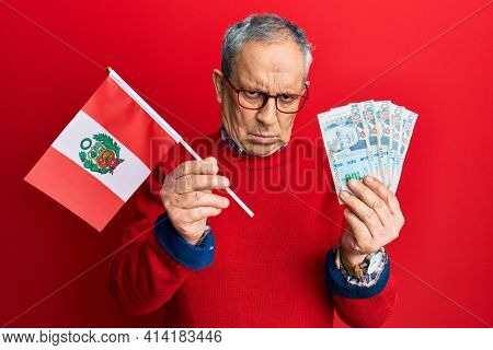 Handsome senior man with grey hair holding peru flag and peruvian sol banknotes skeptic and nervous, frowning upset because of problem. negative person. 