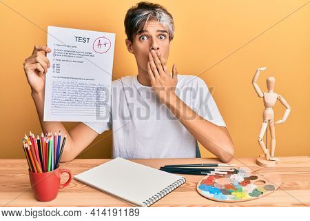 Young hispanic man artist holding passed exam sitting on the table covering mouth with hand, shocked and afraid for mistake. surprised expression 