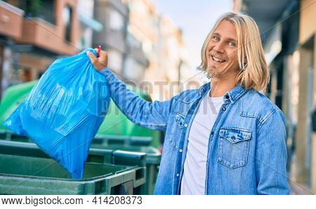 Young blond scandinavian man throwing waste bag to the container at the city.