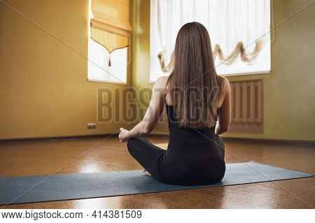 A Woman In Black Sportswear, Practicing Yoga, Is Engaged In Meditation In The Lotus Position On A Gy
