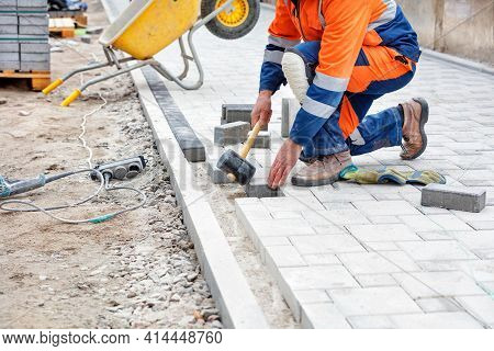A Bricklayer In A Blue And Orange Overalls, Half-squatting, Taps The Paving Slabs With A Black Rubbe