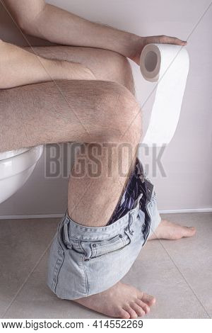 Man Sitting On Toilet Bowl At Home Or Hotel, With Toilet Paper Roll In Hands. Abdominal Stomachache,