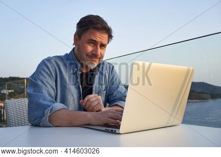 Happy man working online with laptop computer on terrace. Home office, browsing internet, tele working, video chat. Businessman on vacation at sea. Escape, freedom, getaway.