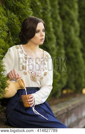 Relaxing Sexy Caucasian Girl In Rural Decorated Dress Pouring Milk In Front Of Line Of Fir-trees