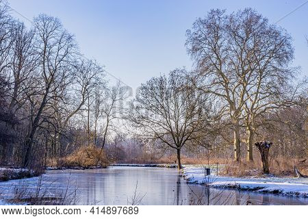 Winter Landscape With A Pond With Its Frozen Waters, Frost And Snow, Surrounded By Huge Bare Trees, 
