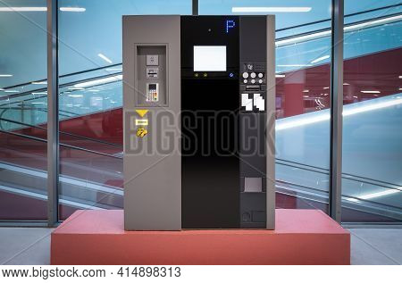 Modern Gray-black Parking Ticket Machine In Parking Garage In Front Of Glass Wall With Escalators In