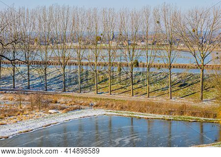 Bare Trees In Line, A Pond With Its Frozen Waters, Snow, Frost With The Juliana Canal And The Maas R