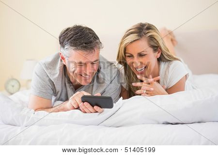 Happy mature couple lying on a bed in a bedroom and laughing watching a mobile phone