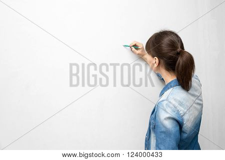 back of young woman in casual clothes with pencil drawing or writing on empty wall