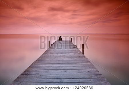 Unrecognizable person sitting on edge of wooden pier at sunset of red and violet colors