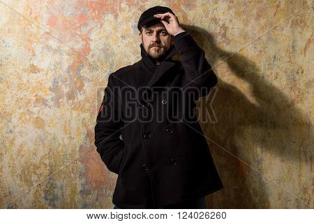 attractive young bearded man in a pea coat and cap is standing at the wall