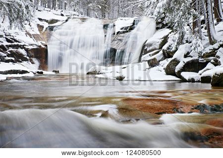 Winter view over snowy boulders to cascade of waterfall. Wavy water level. Stream in deep freeze.