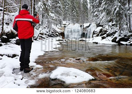 Photograph in red jacket with digital camera in hands is taking photo of winter frozen waterfall