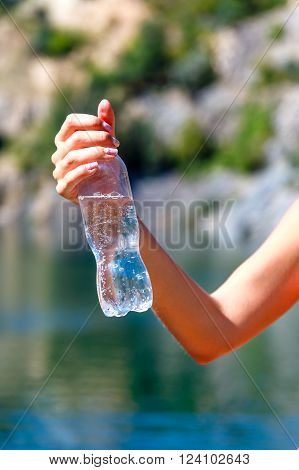 Female hand holding a plastic bottle of mineral water on a background of mountain river at sunny summer day