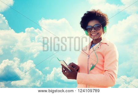 technology, lifestyle and people concept - smiling african american young woman or teenage girl with smartphone and headphones listening to music over blue sky and clouds background