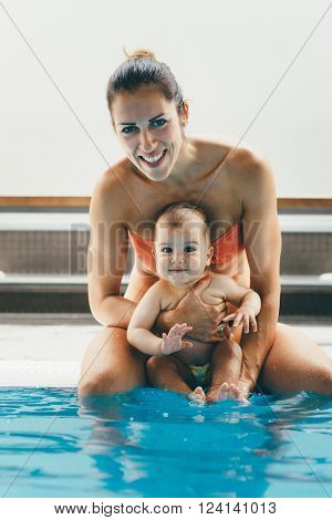Portait of mother with baby boy sitting on the swimming pool edge and splashing. Smiling looking at camera