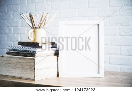 Desk with blank picture frame and pencils in iron mug placed on book and wooden box. Mock up