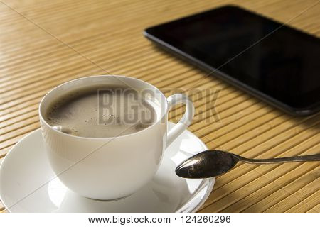 Coffee cup and saucer on a light bamboo table.Tablet computer are near