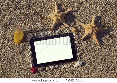 Photo frame made of seashells and pebbles with two starfish and orange juice bottle placed next to it and tablet computer within the frame with blank screen on the beach