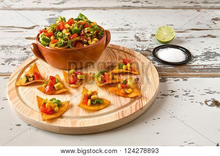 Nachos chips and vegetables in an earthenware bowl on a chopping board and tequila over a wooden table