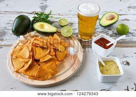 Nachos chips on a chopping board with sauces beer and avocado over an old wooden table