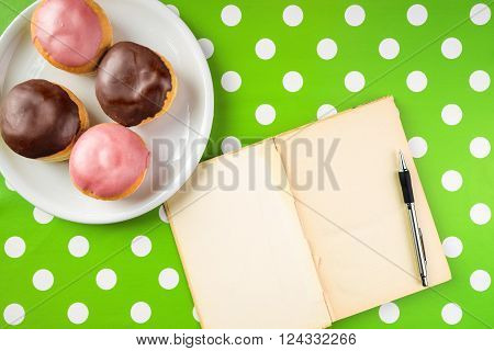 Vintage notebook with recipe for delicious homemade donuts with topping top view of cookbook with blank pages as copy space and tasty doughnuts on a plate over polka dotted surface.