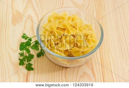 Uncooked bow-tie pasta in glass bowl and sprig of a parsley on a light wooden surface