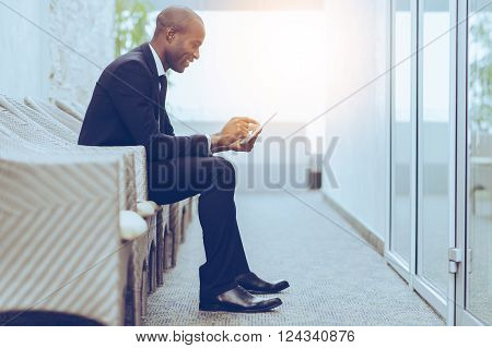 Checking his business schedule. Side view of cheerful young African businessman working on digital tablet while sitting on the chair